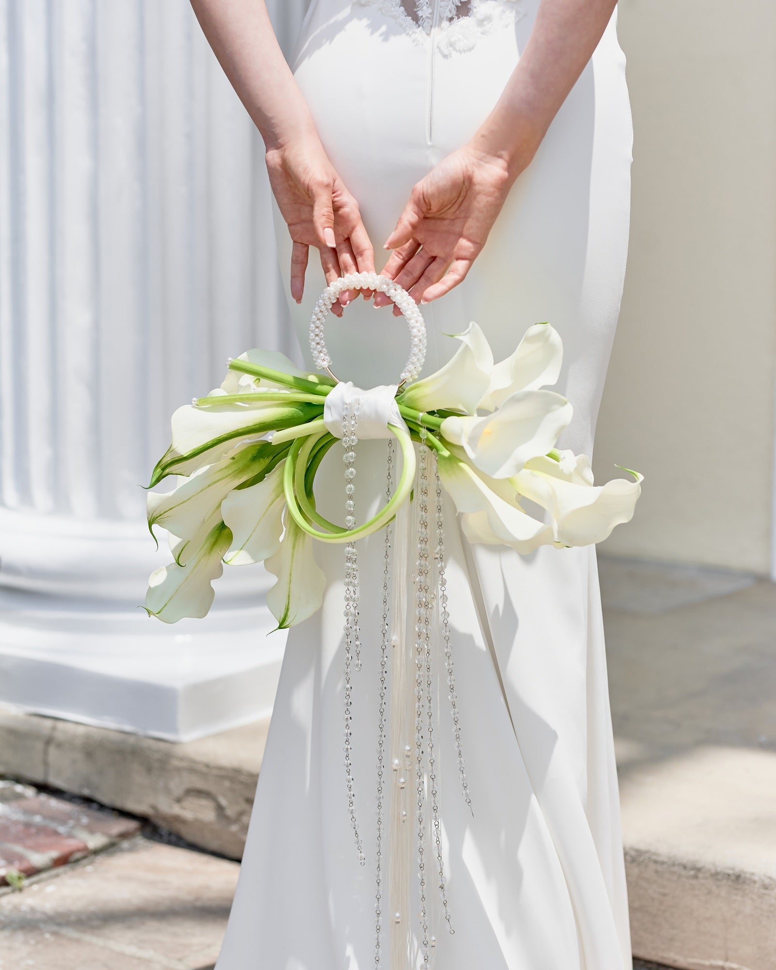 Modern bridal bouquet designed as a sculptural purse with white calla lilies, pearl handle, and crystal tassel chains, held by a bride in an elegant gown.