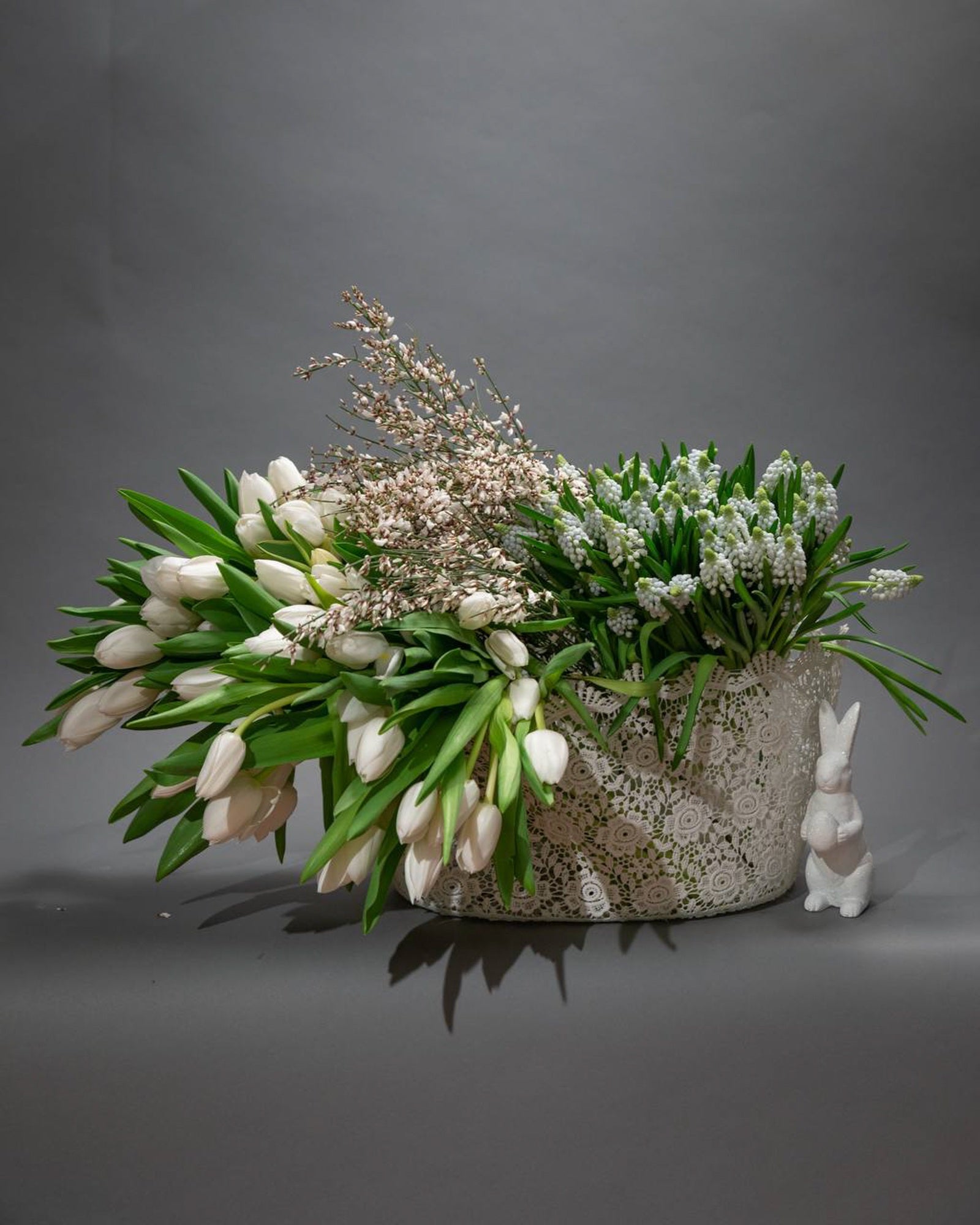 Elegant basket of white tulips, muscari, and waxflower in a white lace-pattern container with a ceramic bunny accent, styled on a neutral background.