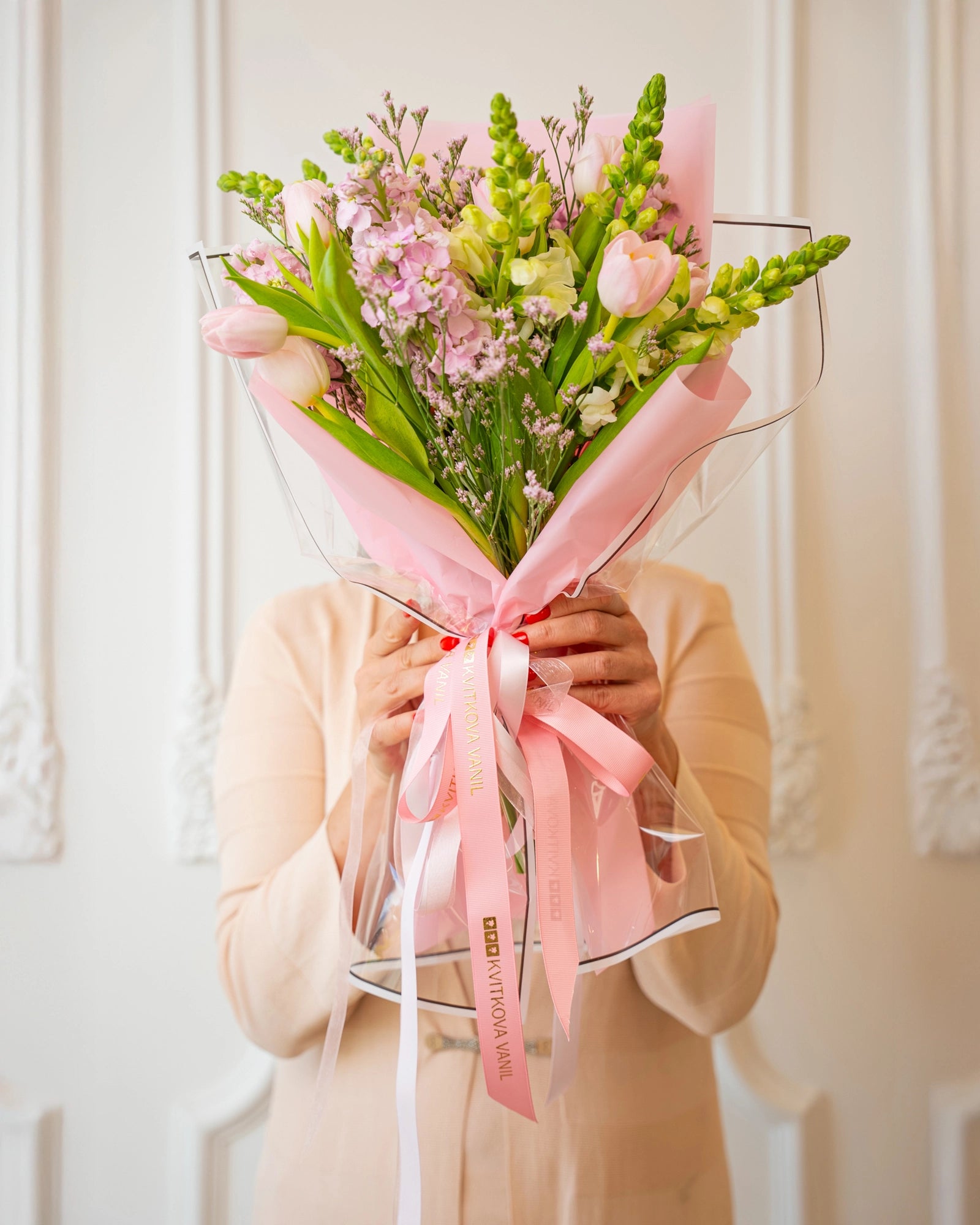 Hand-held view showing the bouquet's pink tulips, cream snapdragons, and branded ribbons, presented for gifting