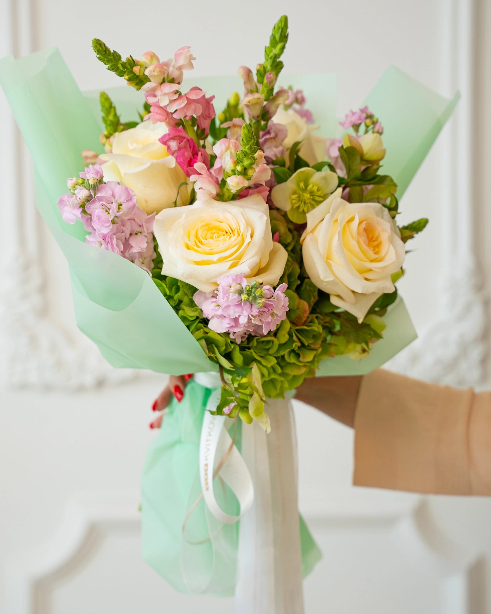Front view of garden-style bouquet with cream roses, pink snapdragons, stock and chartreuse hellebores wrapped in mint paper